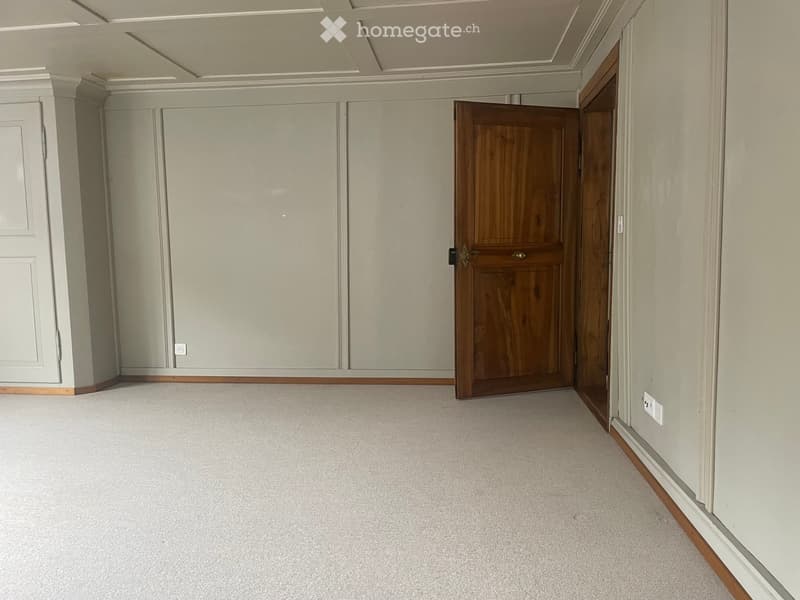 Empty room with light grey walls, carpeted floor, and a wooden door leading to another space.