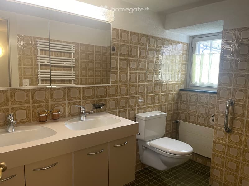 Bathroom with double sink, toilet, mirror, radiator, and window. Brown tiled walls and beige cabinetry.