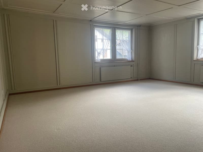 Empty room with beige carpet, light grey walls, and large windows with white curtains. Radiator visible under one window.