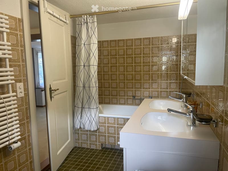 Bathroom with beige tiles, double sink, bathtub, mirror, radiator, and a partially open door leading to another room.