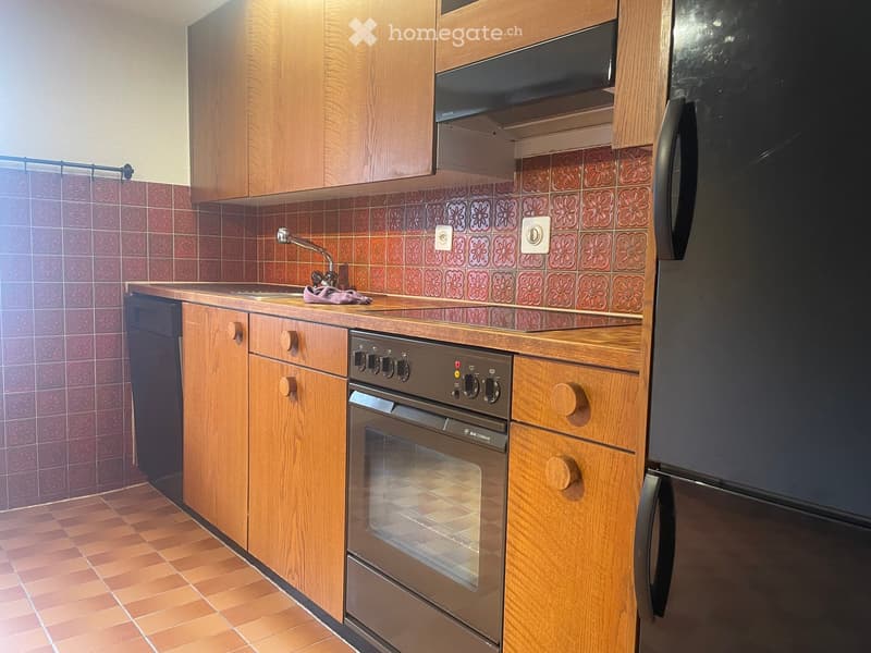 A cozy kitchen with wooden cabinets, a stainless steel oven, a refrigerator, and a sink with a faucet. The walls are tiled with a floral pattern, and the floor is tiled in a warm, earthy tone.