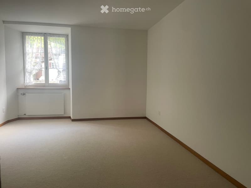 Empty room with beige walls, carpeted floor, and a single window with white curtains. A radiator is mounted below the window.