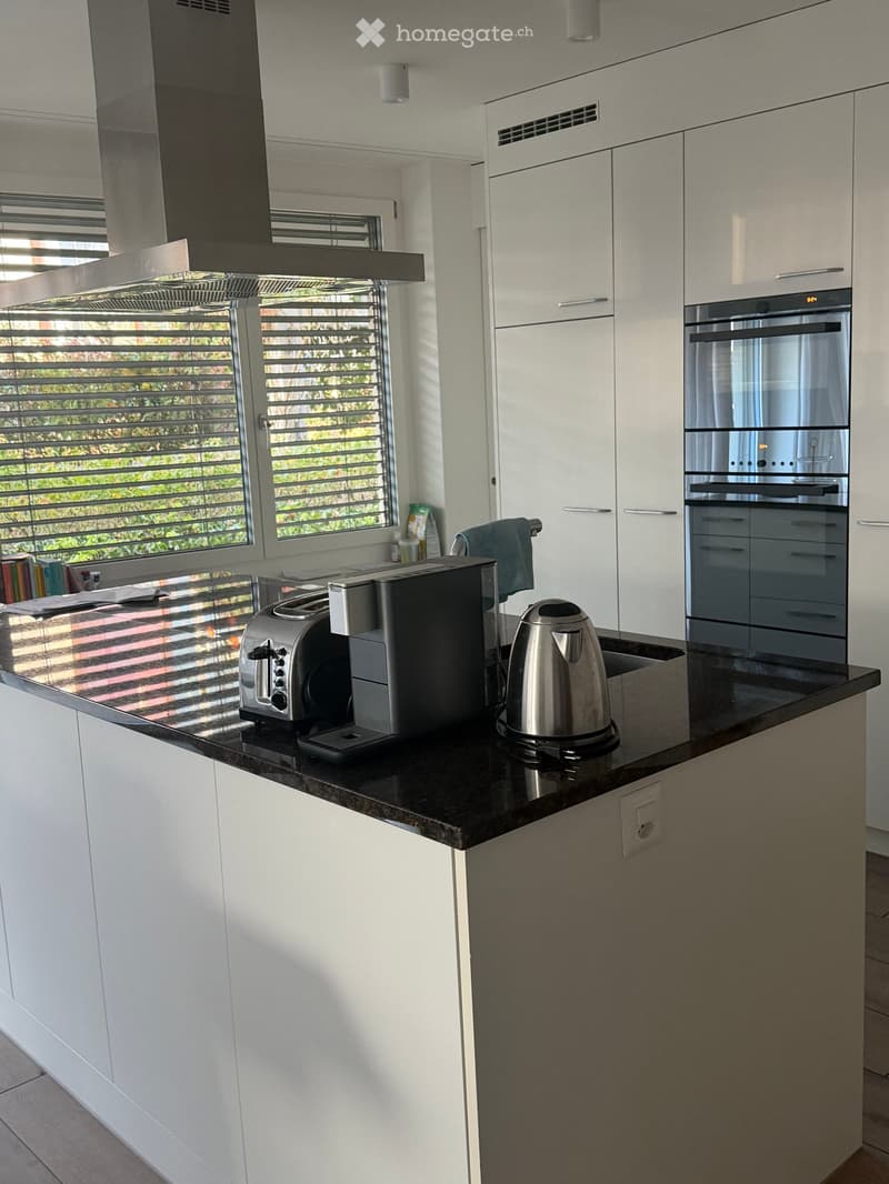 Modern kitchen with white cabinets, black countertop, stainless steel appliances including a coffee maker, toaster, kettle, and oven. Large windows with blinds allow natural light to enter.