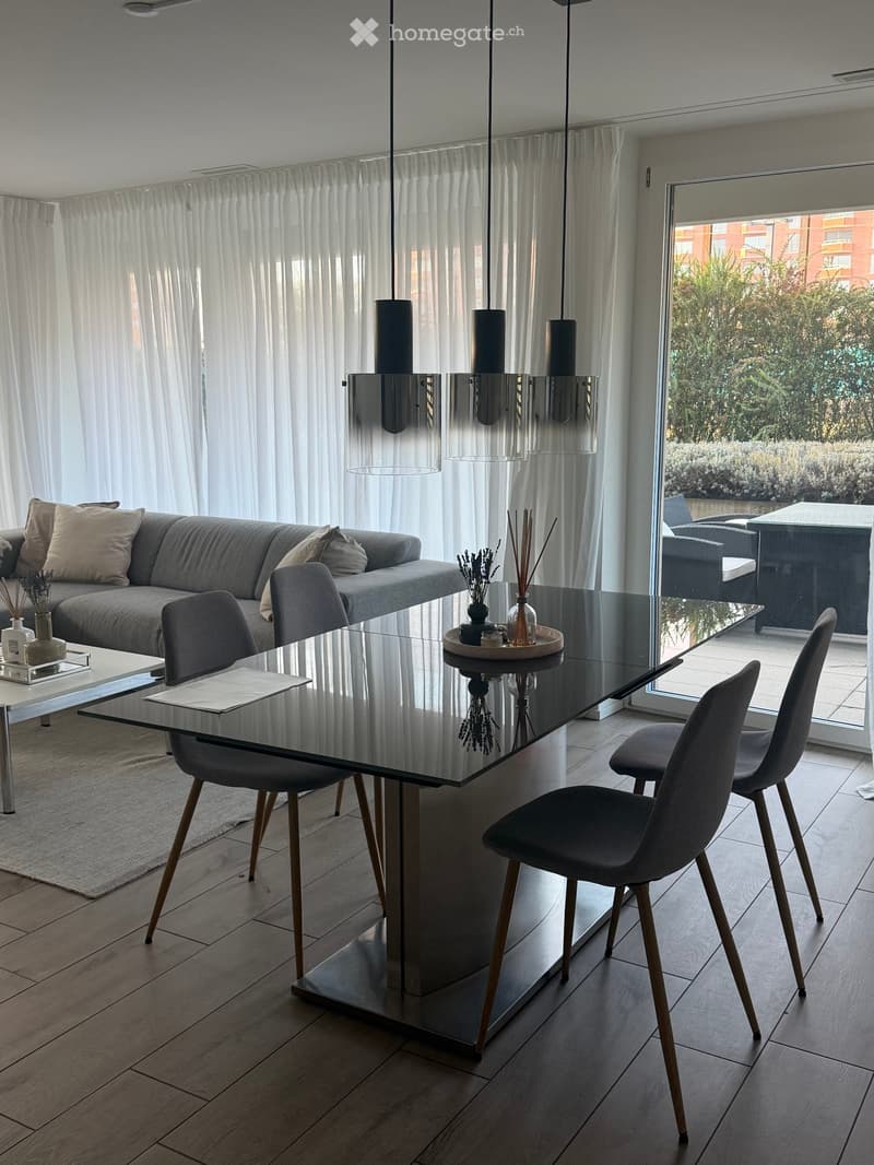 Modern living and dining area with a grey sofa, glass coffee table, dining table with chairs, decorative items, and pendant lights. Large windows with white curtains lead to a balcony.