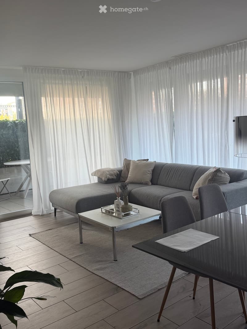 Modern living room with grey sectional sofa, white coffee table, side table, decorative vase, and a plant. Large windows with sheer curtains and wooden flooring.