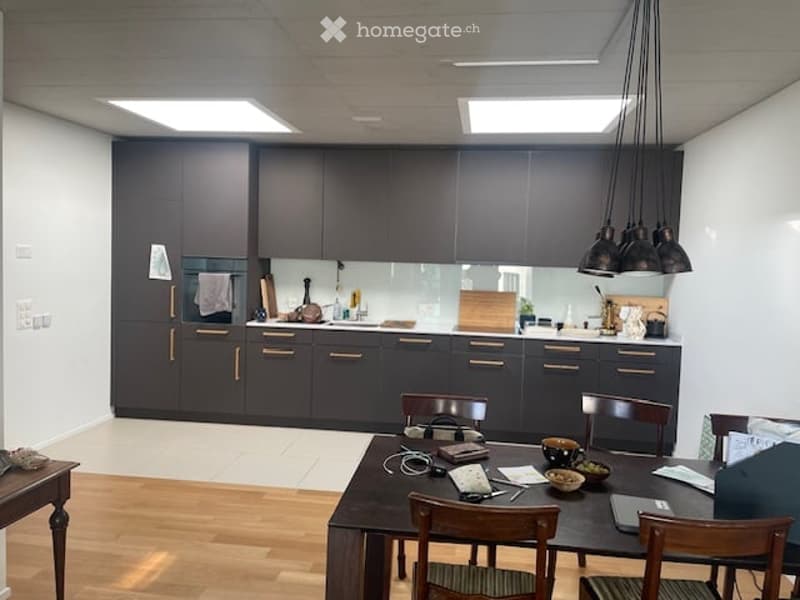 Modern kitchen with dark grey cabinets, a central island, and a dining area with a wooden table and chairs. The kitchen features a sink, faucet, and various small appliances and items on the counter.