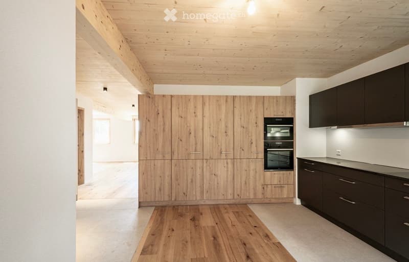 Modern kitchen with wooden cabinets, dark lower cabinets, and a built-in oven. The space features exposed wooden beams on the ceiling and a light wooden floor.