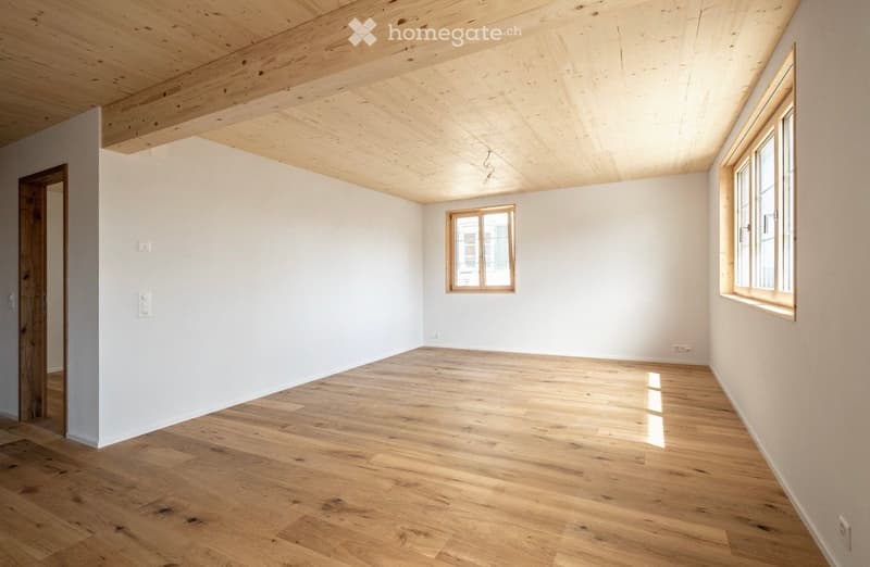 Empty room with wooden flooring, white walls, and exposed wooden ceiling beams. Two windows and a door are visible.