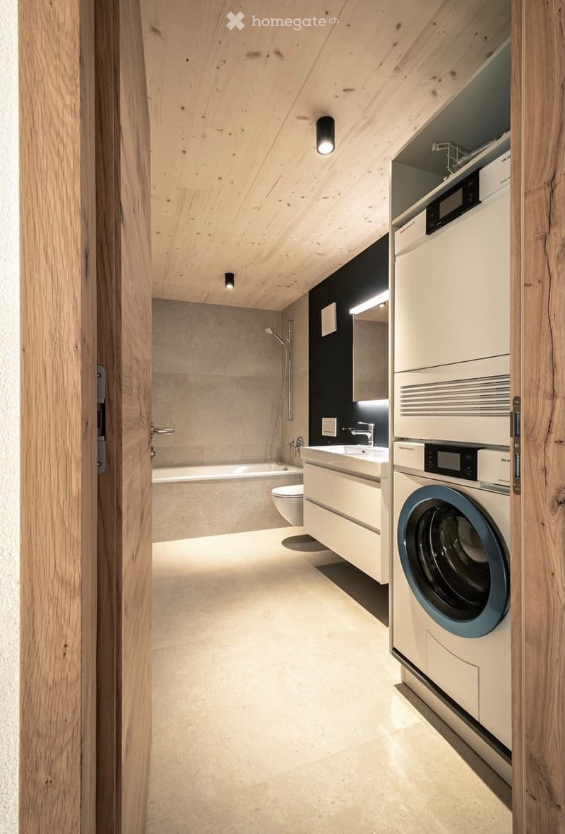 Modern bathroom with a bathtub, toilet, sink, and washing machine. Features wooden ceiling and walls, sleek fixtures, and a minimalist design.