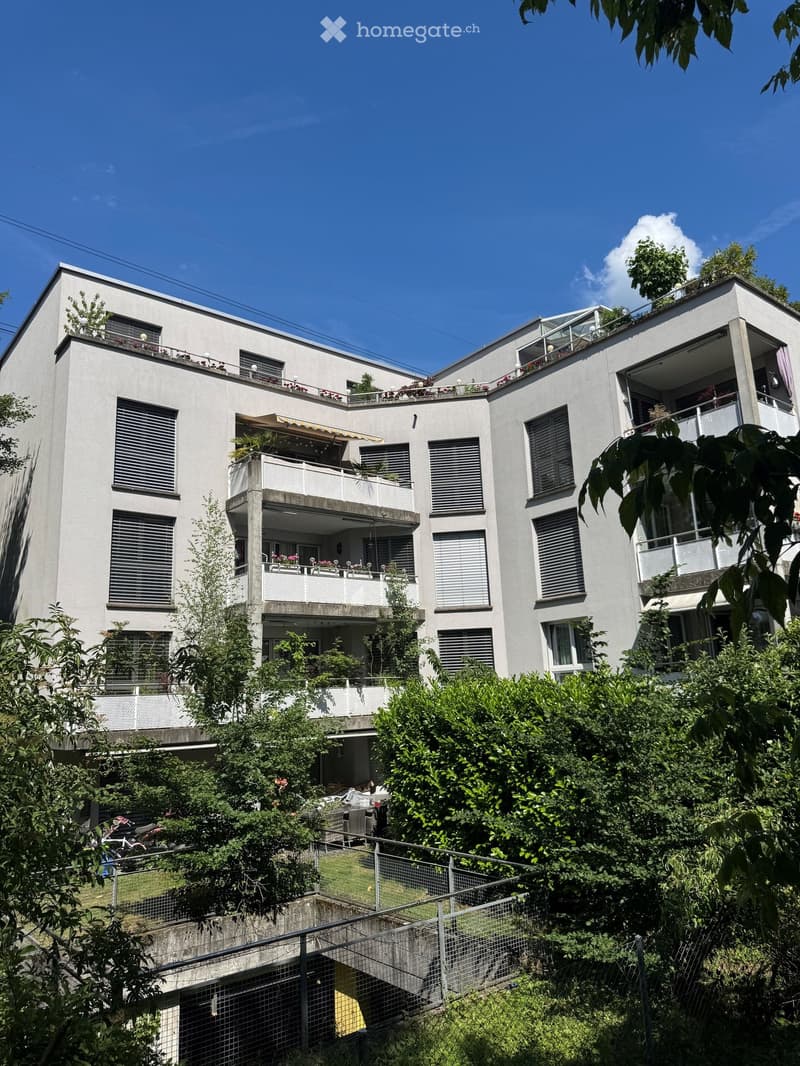 Modern multi-story residential building with balconies, large windows, and greenery on the roof and surrounding area.