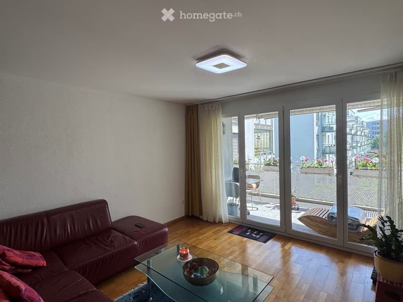 Modern living room with wooden flooring, large sliding glass doors leading to a balcony, a red leather sofa, a glass coffee table with a bowl, and a potted plant near the window.
