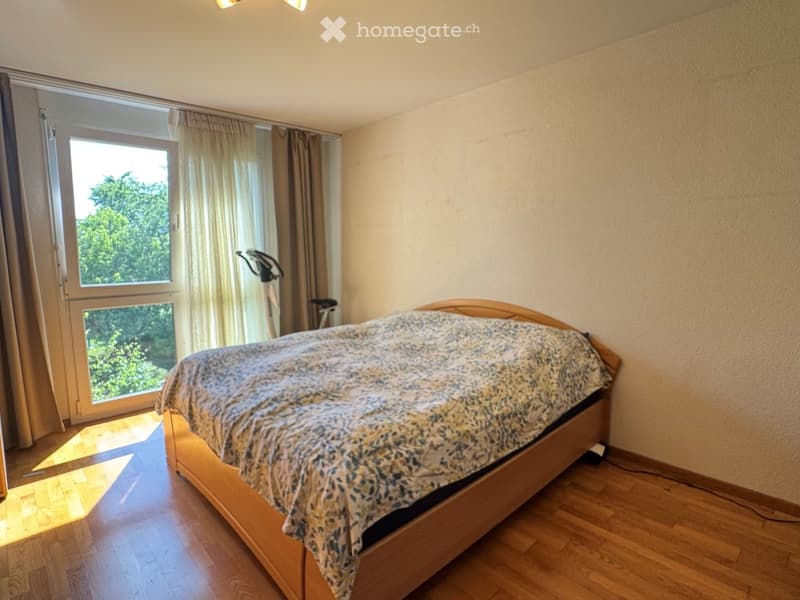 Bright bedroom with wooden flooring, large window with curtains, bed with patterned bedspread, and exercise bike in the corner.