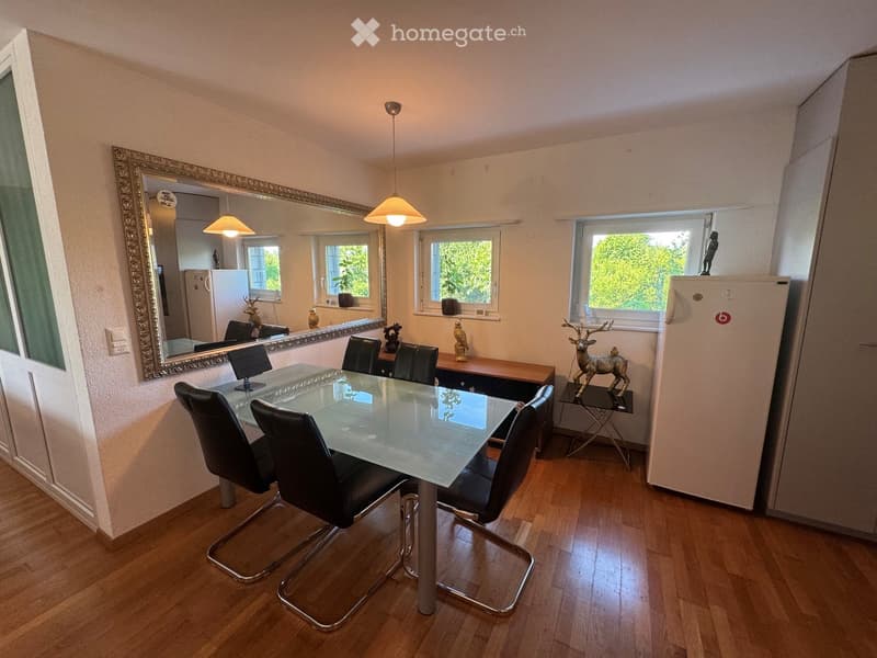 Dining room with a glass table, black chairs, a large mirror, and a refrigerator. Natural light from multiple windows illuminates the space.