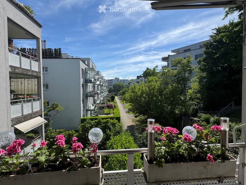 Balcony with flower boxes containing pink flowers and decorative glass orbs, overlooking a landscaped area with trees and modern apartment buildings.