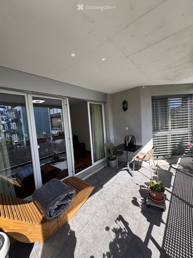 Balcony with tiled floor, wooden bench, small table with chairs, potted plant, and a fryer. Large sliding glass doors lead to the interior.