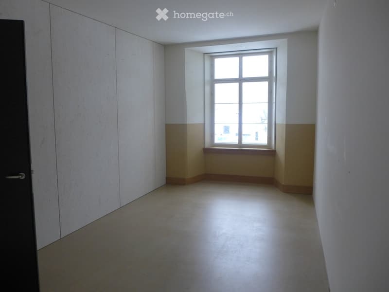 Empty room with white walls, a window with multiple panes, and a simple wooden sill. The floor is light-colored and the room has a door on the left side.