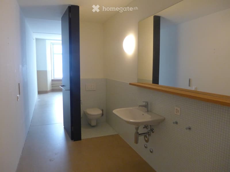 Modern bathroom with a toilet, sink, and mirror. The walls are tiled with a light grey pattern, and the floor is a light brown tile. A round light fixture is mounted on the wall above the mirror.