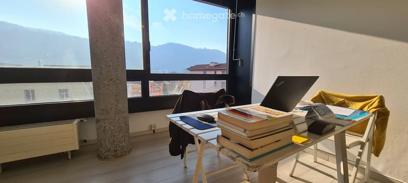 Office room with a desk, books, laptop, and a heater. The room has large windows with a scenic view of mountains and buildings.