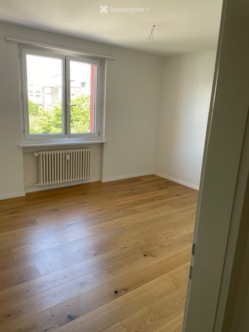 Empty room with wooden flooring, white walls, and a window with a radiator below it.
