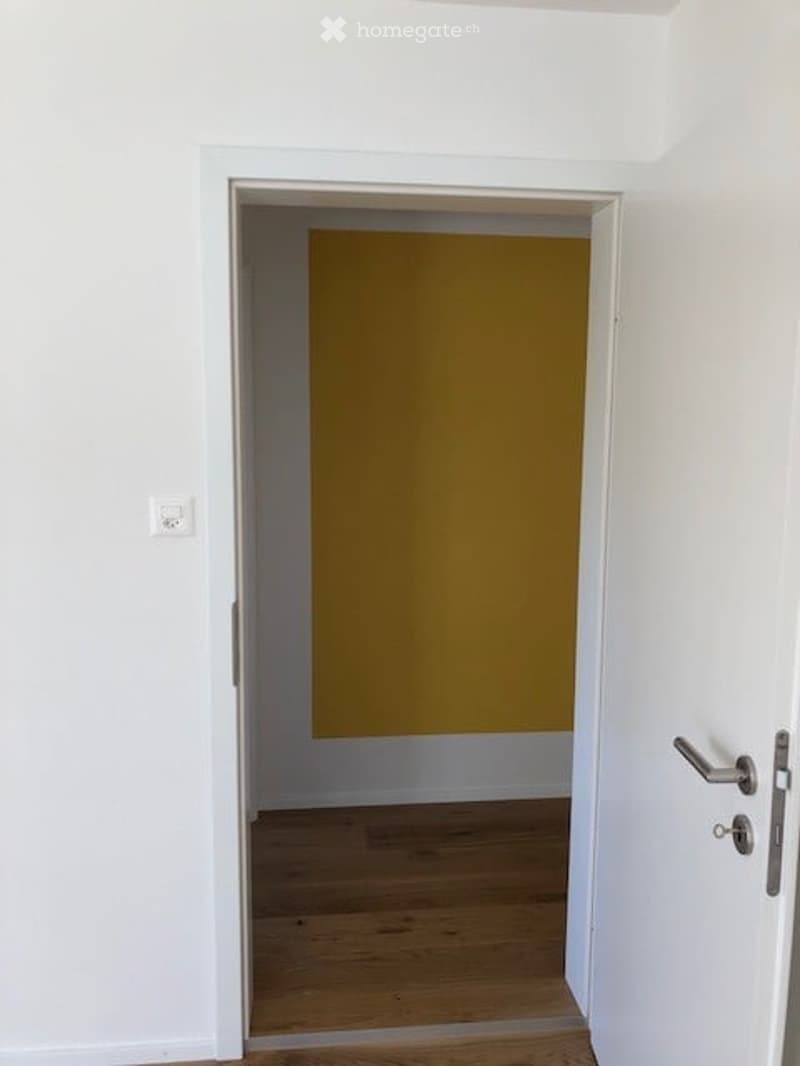 A narrow hallway with white walls and wooden flooring, leading to a room with a yellow accent wall.