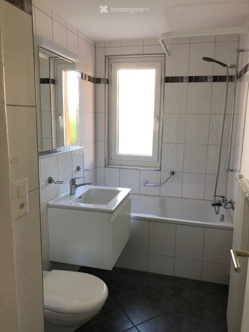 Modern bathroom with white tiles, a sink with a modern faucet, a bathtub, a toilet, and a showerhead. The room features a window for natural light and a mirror above the sink.