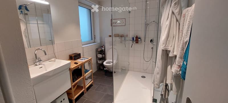 Modern bathroom with white tiles, a sink, toilet, shower, and a small wooden table holding a speaker and other items. Towels and a bathrobe are hanging on the wall.