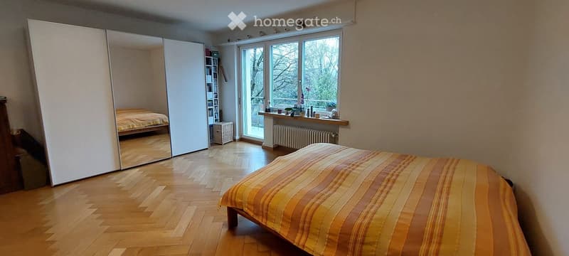 Bright bedroom with wooden flooring, large window with view, bed with striped bedding, large mirror, bookshelf, and small table near the window.