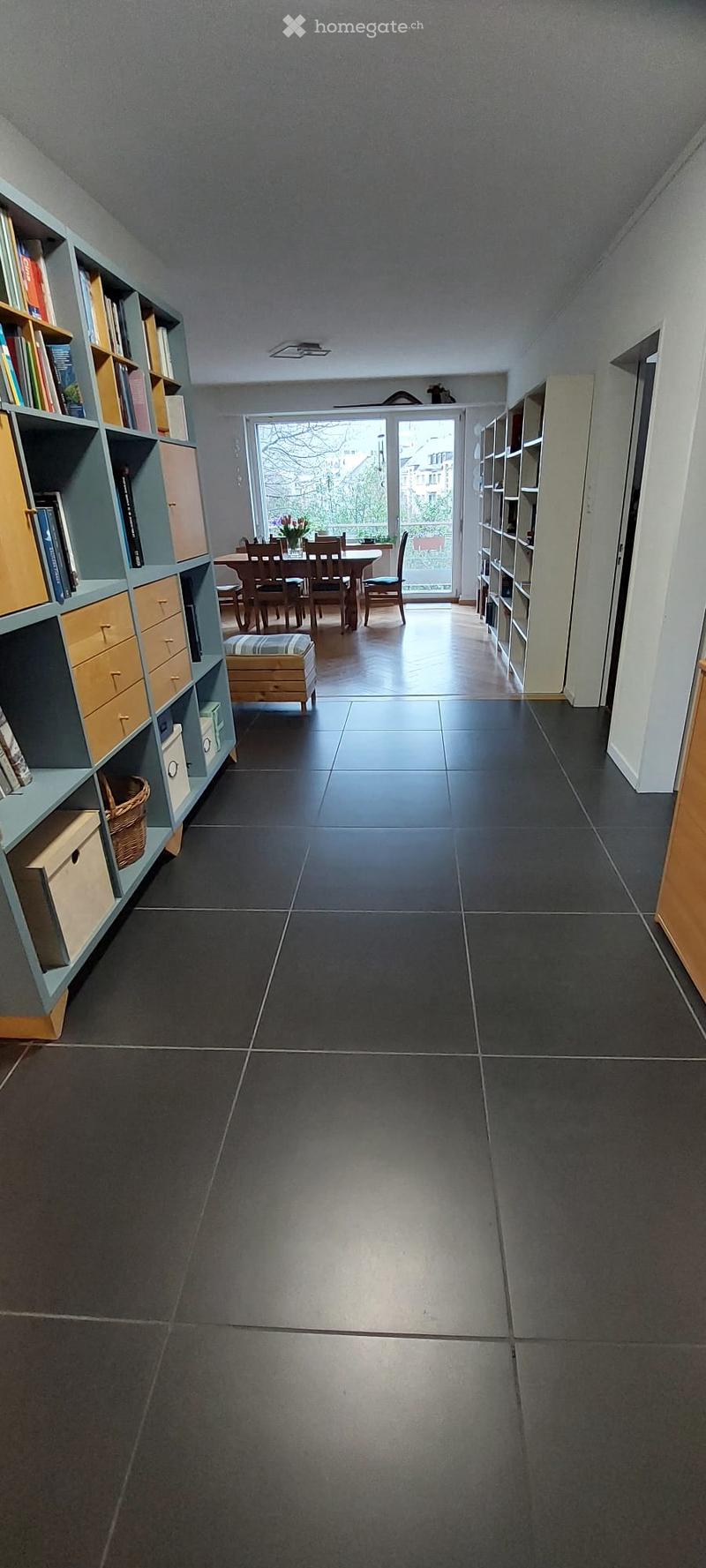 Hallway with dark tiled floor, built-in bookshelves on both sides, leading to a dining area with a table and chairs, and a view through a large glass door to an outdoor area.