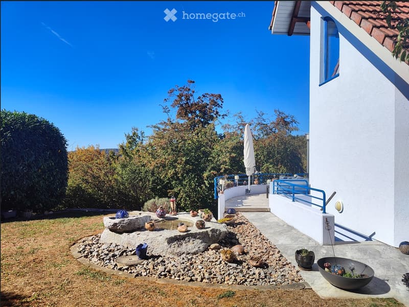 Garden area with a stone fountain, decorative bowls, and a small patio with a blue railing, adjacent to a house with a tiled roof and white walls.
