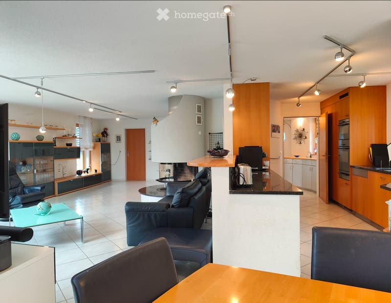 Modern living room with a black leather sofa, glass coffee table, wooden dining table, and a small kitchen area with cabinets and appliances.