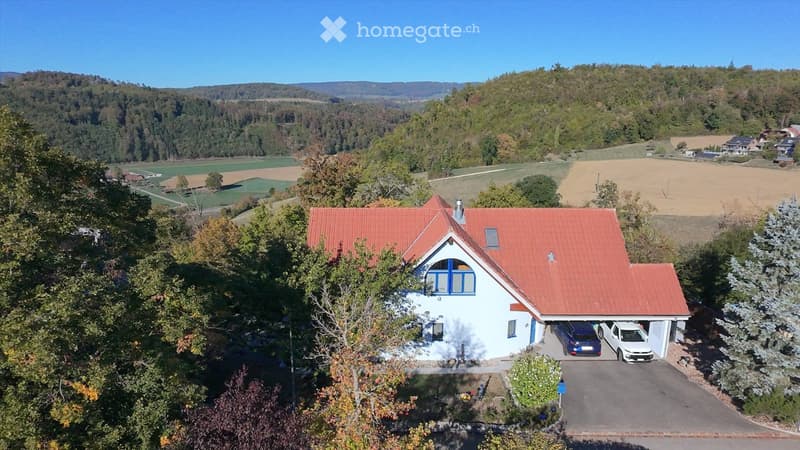 Aerial view of a two-story house with a red-tiled roof, white walls, and blue window frames. The house features a garage with two parked cars and is surrounded by a landscaped garden. The background shows a scenic countryside with fields, trees, and dista