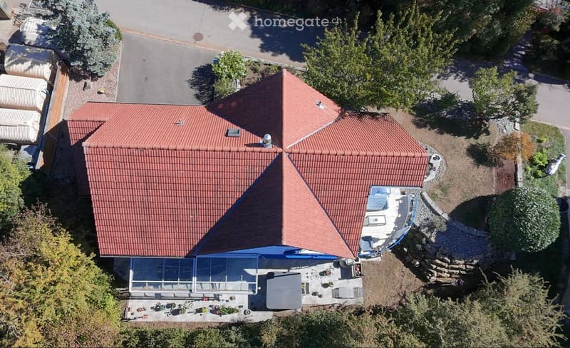 Aerial view of a two-story house with a red-tiled roof, surrounded by trees and a landscaped garden. The house features a covered patio area with outdoor furniture and a small swimming pool.