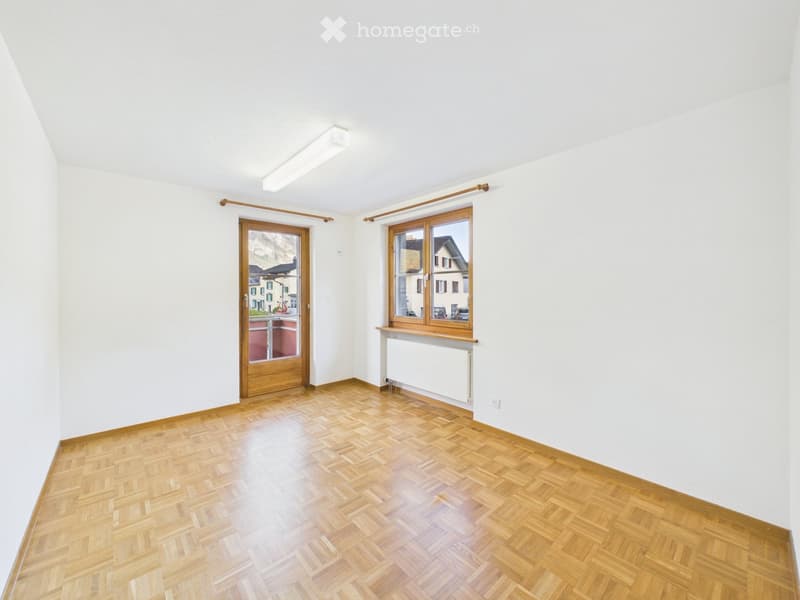 Empty room with wooden herringbone flooring, white walls, large window with wooden frame, and a door leading to a balcony.
