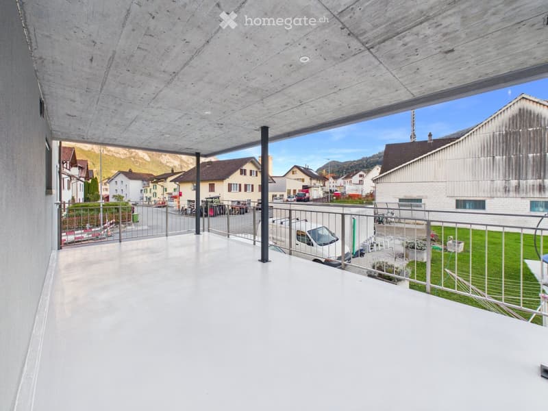 Modern balcony with white concrete floor, metal railing, and a view of a town with houses, mountains, and parked vehicles.