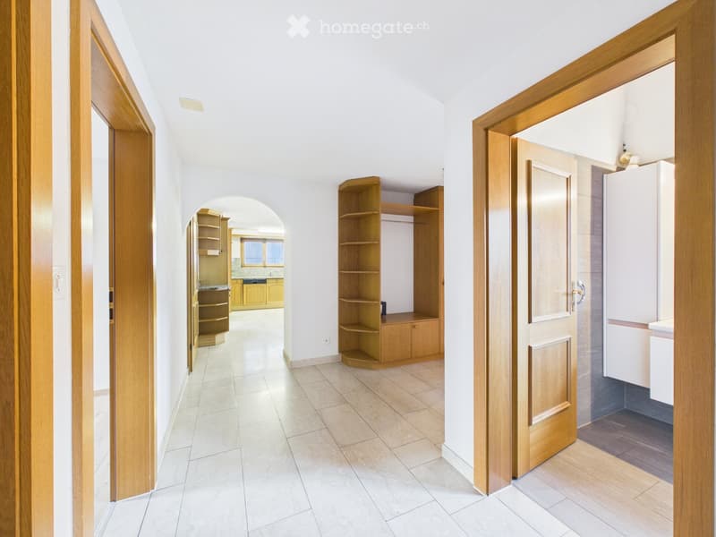 Bright hallway with tiled floor, wooden doors, and built-in wooden shelving unit. The hallway leads to a kitchen area with cabinets and a refrigerator visible in the background.