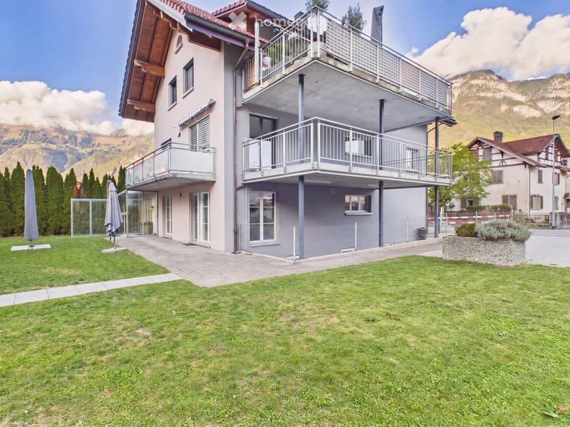 Modern two-story house with balconies, surrounded by a well-maintained lawn and trees, with mountain views in the background.