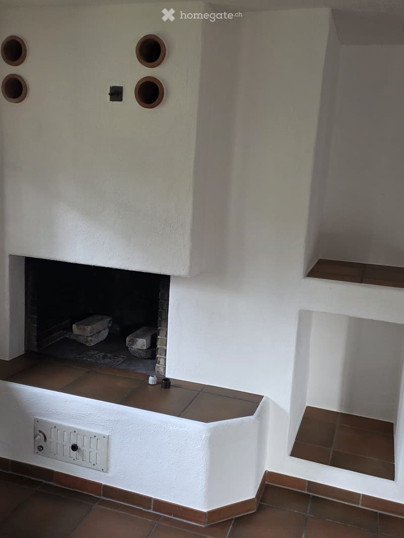 A hallway with a built-in fireplace featuring a white plaster surround and brown tiles. The fireplace has four circular openings above and a small electrical outlet below.