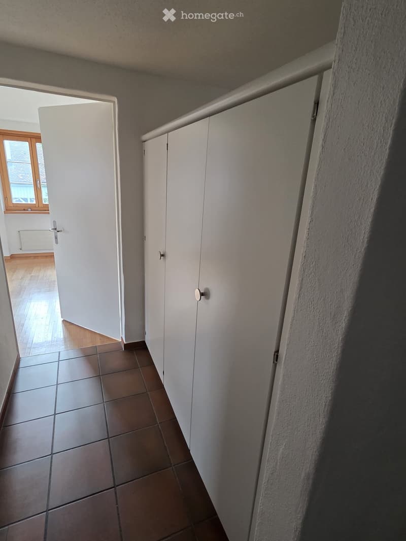 A narrow hallway with tiled flooring, white built-in cabinets, and an open doorway leading to another room with wooden flooring and a window.