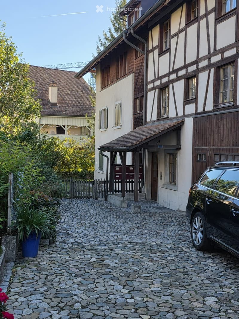 A traditional half-timbered house with a cobblestone pathway leading to its entrance, featuring a small porch and a parked car nearby.