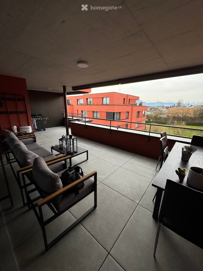 Modern balcony with grey sofa, wooden-framed chairs, small table, lanterns, and a view of a red building and mountains.