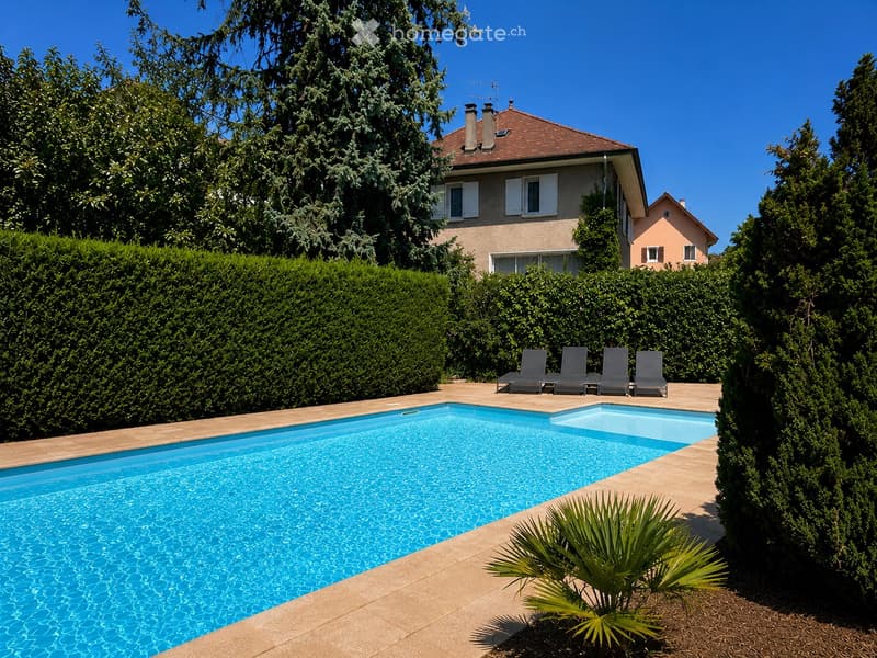 A rectangular swimming pool with clear blue water, surrounded by a tiled deck, lounge chairs, and lush greenery including tall trees and shrubs.