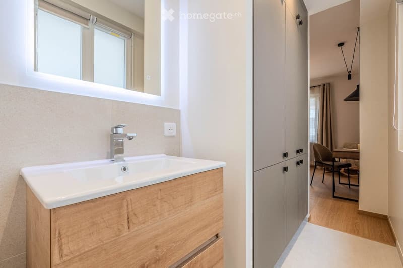 Modern bathroom with a white sink, wooden cabinet, and a mirror above it. The adjacent room features a wooden floor, a chair, and a small table.