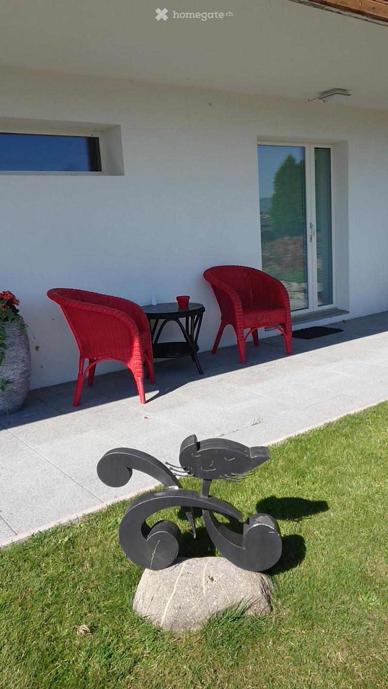 Outdoor patio area with two red wicker chairs, a small black table, a decorative vase with flowers, and a modern metal sculpture on a rock in the grass.