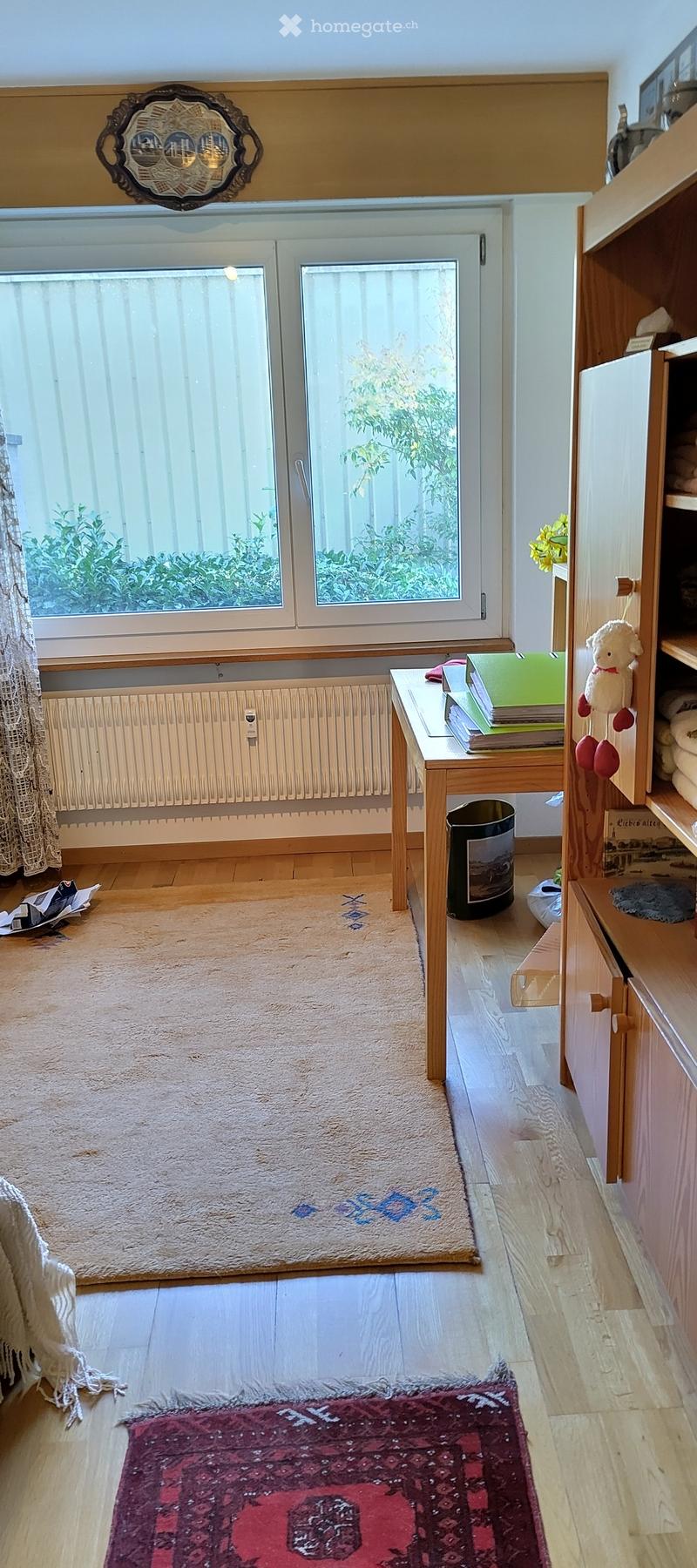 A cozy, well-lit room with wooden flooring, two rugs, a small table with books and a toy, a radiator, and a window with a view of greenery.