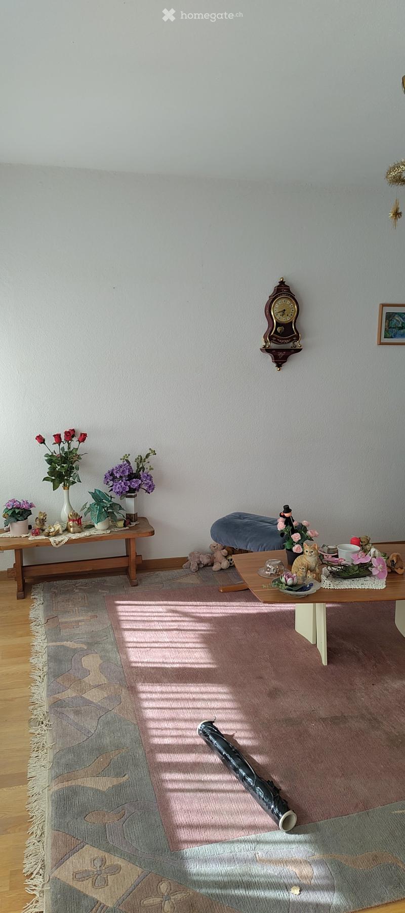 A cozy living room with wooden floors, a patterned rug, a small wooden table with vases of flowers, a blue upholstered stool, and a wall-mounted clock.
