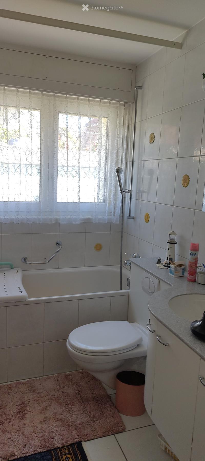 Bathroom with a bathtub, toilet, sink, and cabinet. Features a window with a curtain, a handrail, and a trash can.