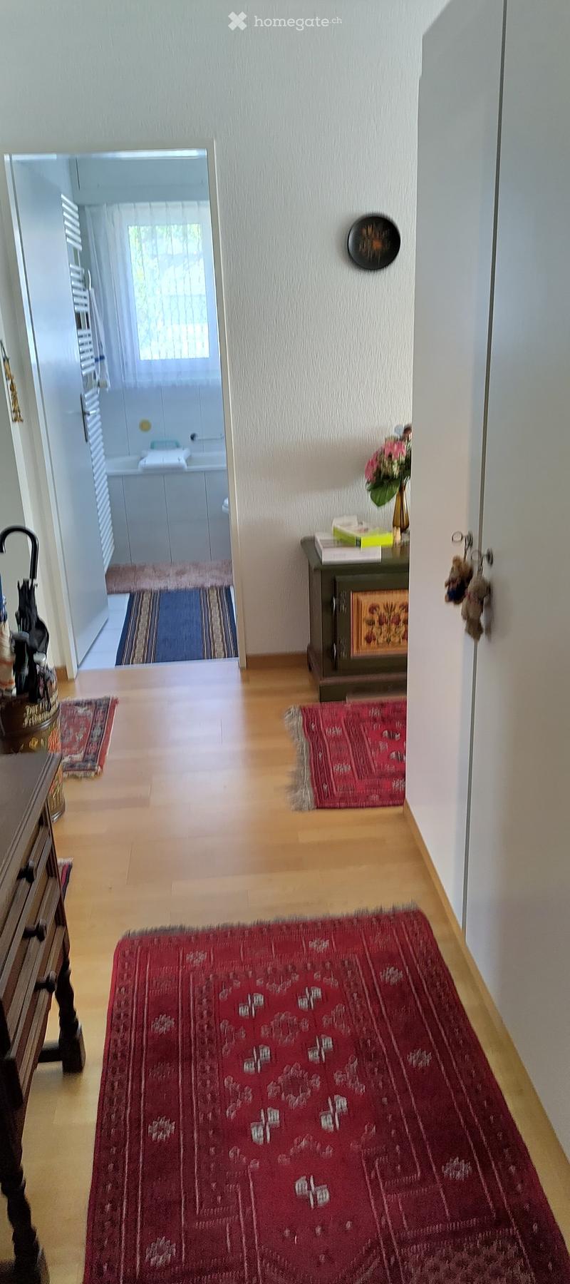 A well-lit hallway with wooden flooring, red rugs, and a small table with a vase of flowers. A doorway leads to a bathroom with white cabinets and a sink.