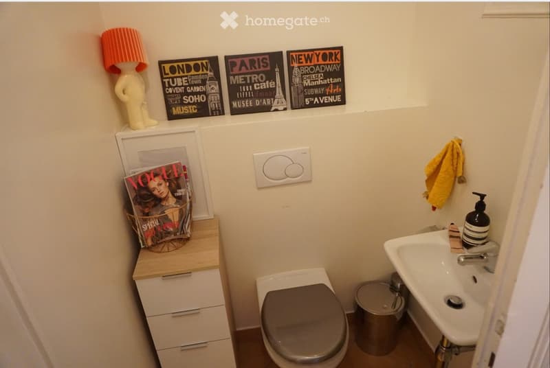 A compact bathroom with a toilet, sink, and cabinet. The sink has a modern faucet and a soap dispenser. A small cabinet holds magazines and a decorative lamp. A towel hangs on the wall, and a trash bin is placed near the toilet.