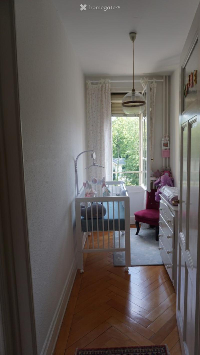 A small, well-lit room with wooden flooring, a crib, a red chair, a dresser, and toys visible. The room has a view of a balcony through an open door.