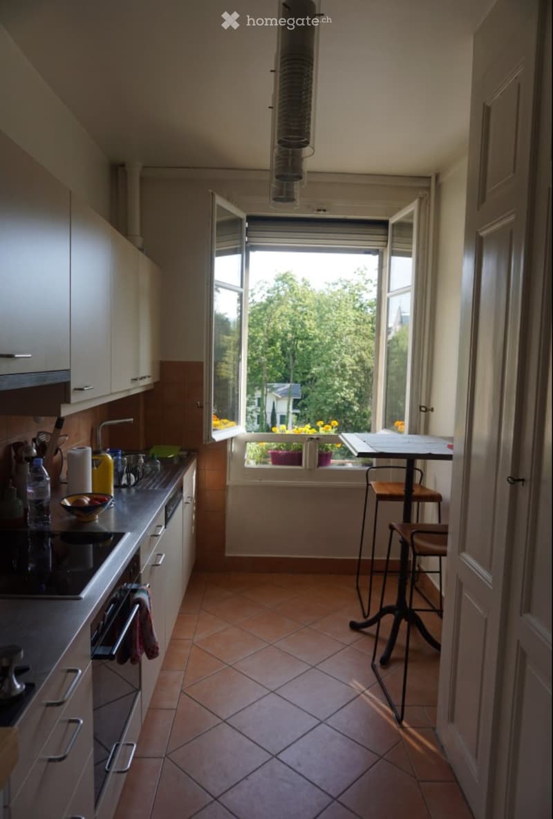 Modern kitchen with white cabinets, stainless steel appliances, and a small dining area with a table and stools. The room features terracotta tiles and large windows with a view of greenery.